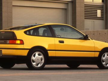 Un Honda CR-X holandés amarillo brillante con portón trasero de los años 80 está aparcado fuera de un edificio urbano con las puertas del garaje cerradas.