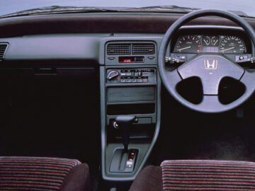 Intérieur d'une Honda CR-X avec un volant portant le logo emblématique de Honda, un tableau de bord et un panneau de commande. Les sièges sont en tissu rayé, tandis que le levier de vitesse est placé en évidence sur la console centrale. Ce modèle néerlandais allie style et fonctionnalité.