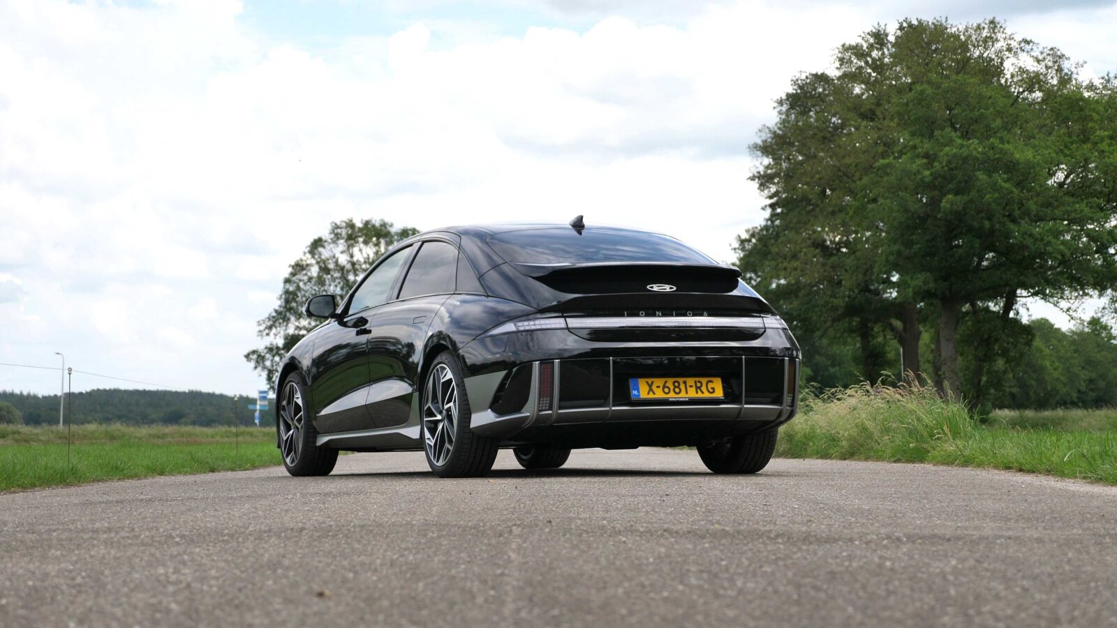 Een zwarte Hyundai IONIQ 6 met kenteken "X-681-RG" staat geparkeerd op een landelijke weg, omgeven door groene bomen en gras onder een gedeeltelijk bewolkte hemel. De auto beschikt over een actieradius van 600 kilometer, perfect voor lange ritten door schilderachtige landschappen.