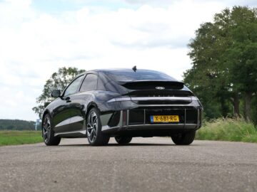 Een zwarte Hyundai IONIQ 6 met kenteken "X-681-RG" staat geparkeerd op een landelijke weg, omgeven door groene bomen en gras onder een gedeeltelijk bewolkte hemel. De auto beschikt over een actieradius van 600 kilometer, perfect voor lange ritten door schilderachtige landschappen.