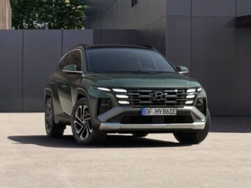 Een groene Hyundai Tuscon staat geparkeerd buiten een modern gebouw met een gevel van reflecterend glas en metaal. Op de kentekenplaat aan de voorkant van het voertuig staat 'OF HY 861E', wat de strakke lijnen van de recente facelift benadrukt.