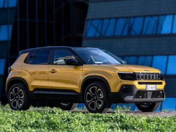 Een gele compacte SUV Jeep Avenger staat geparkeerd voor een modern glazen gebouw met gras op de voorgrond, een bewijs van zijn populariteit (100.000 keer verkocht).