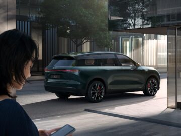 A woman using her smartphone stands on a street near a modern green SUV parked in an urban setting with glass buildings and trees.