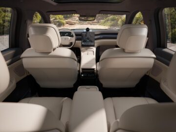 Binnenaanzicht van een moderne SUV met beige lederen stoelen, grote ramen, een schuifdak en een touchscreen-navigatiesysteem op het dashboard. Het voertuig staat geparkeerd op een ruig buitenpad.