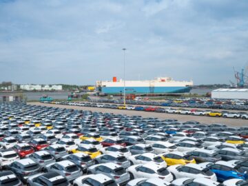 Un grand parking rempli de nouvelles voitures Nissan Qashqai se trouve à proximité d'un cargo à quai. Des grues et des bâtiments industriels sont visibles à l'arrière-plan, créant une image inextricablement liée au port animé de Sunderland. Le ciel est partiellement nuageux.