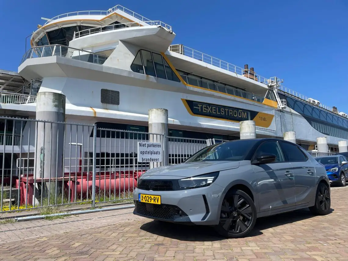 Een grijze auto, mogelijk een Opel Corsa Electric, staat geparkeerd bij een groot schip met de naam "Texelstroom". Het schip is aangemeerd en de auto staat op een met stenen geplaveid terrein voor een metalen hek. Iets verderop draagt een andere compacte EV (een blauwe) bij aan het serene tafereel. Perfect voor een lange roadtrip.