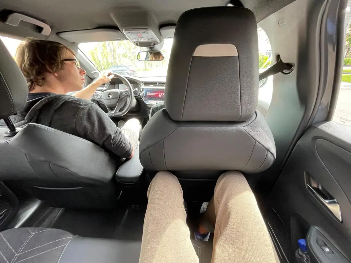 View from the back seat of a car shows a driver in a black hoodie driving the Opel Corsa Electric, with beige pants visible from the perspective of the passenger in the back seat. This compact EV is perfect for a long road trip and makes for an eco-friendly journey.