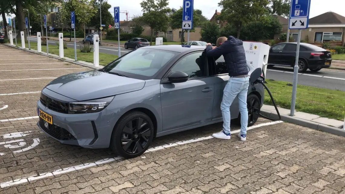 Een persoon laadt zijn lichtgrijze Opel Corsa Electric op bij een laadstation voor elektrische voertuigen op een parkeerplaats.