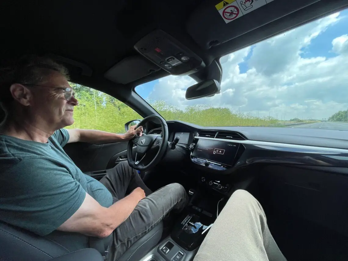 A man drives an Opel Corsa Electric on an open road under partly cloudy skies. The dashboard and interior of the compact EV are visible. Someone else's legs can be seen on the passenger seat, enjoying the road trip.