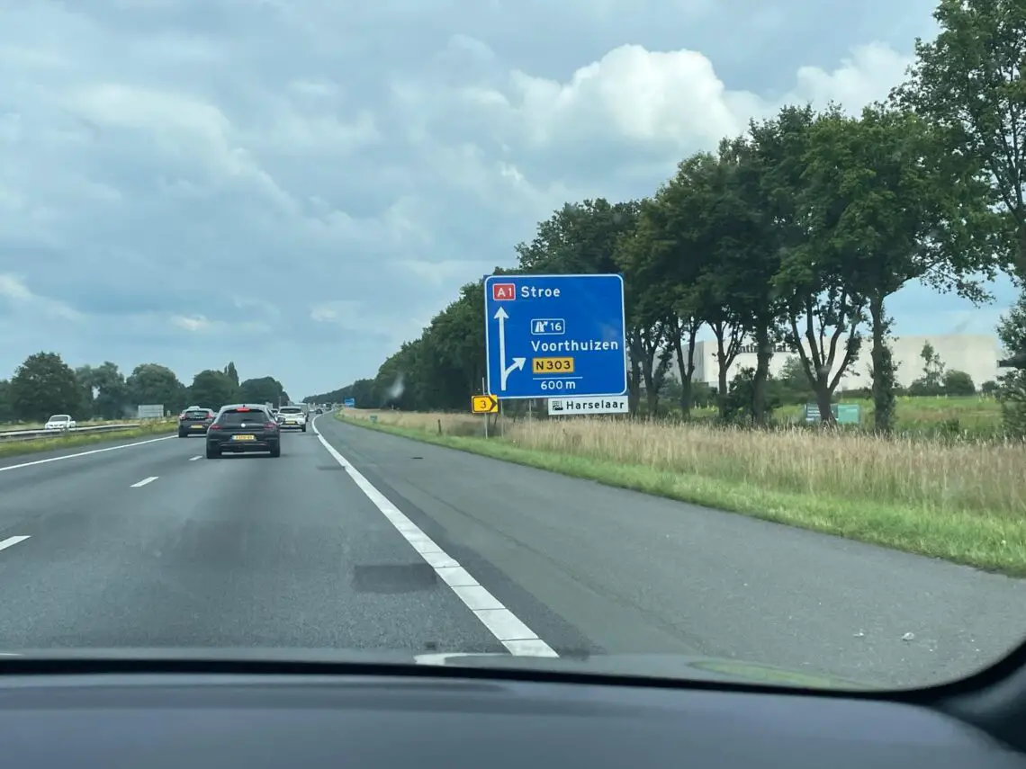 Eine Autobahnansicht mit fahrenden Fahrzeugen, darunter ein Opel Corsa Electric. Ein blaues Schild weist auf die Ausfahrten Stroë und Harselaar in 600 Metern in Richtung N303 hin. Bäume säumen die Straße und der Himmel ist bedeckt: eine ideale Kulisse für einen ruhigen Road Trip.