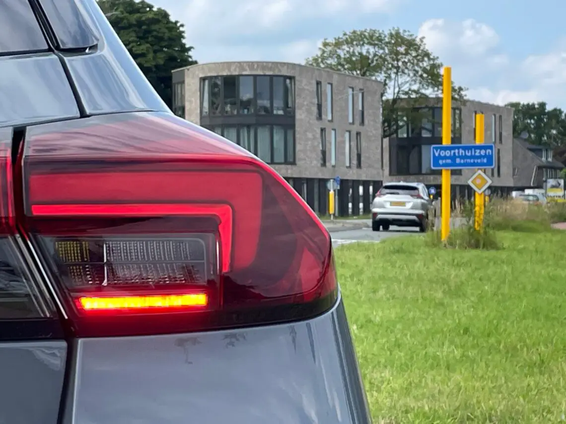 Close-up van het achterlicht van een Opel Corsa Electric met een modern gebouw en een verkeersbord voor Voorthuizen, gemeente Barneveld op de achtergrond op een heldere dag.