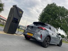 Een grijze Opel Corsa Electric is buiten aangesloten op een zwart laadstation voor elektrische voertuigen, met een grote boom en gebouwen zichtbaar op de achtergrond, klaar voor zijn volgende compacte EV-avontuur.