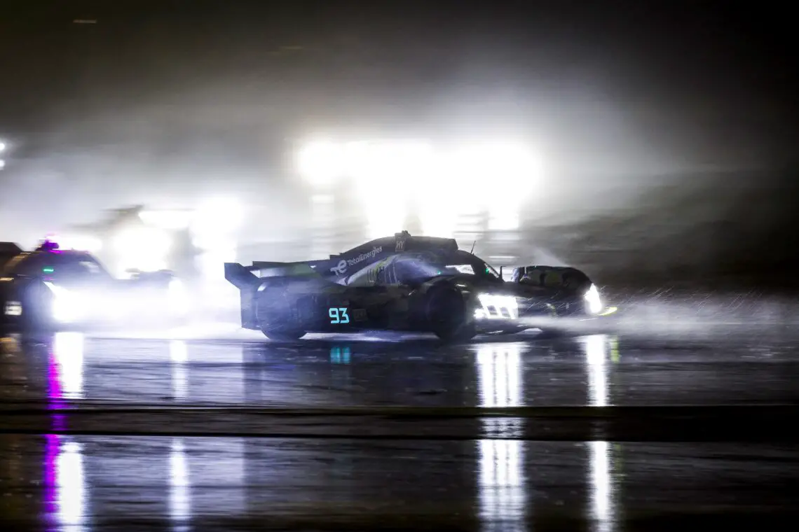 Een Peugeot 9X8-raceauto met nummer 93 rijdt 's nachts door de regen op een natte, reflecterende baan, terwijl felle koplampen van andere auto's het tafereel verlichten.