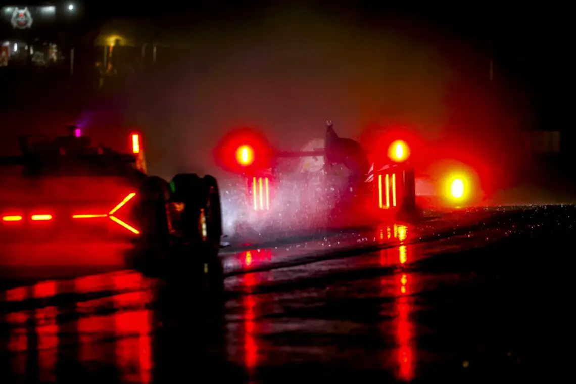 Raceauto's met verlichte achterlichten navigeren op een natte, donkere baan tijdens de 24 uur van Le Mans en creëren reflecties op het oppervlak. Dit is een spektakel dat je minstens één keer in je leven moet meemaken.