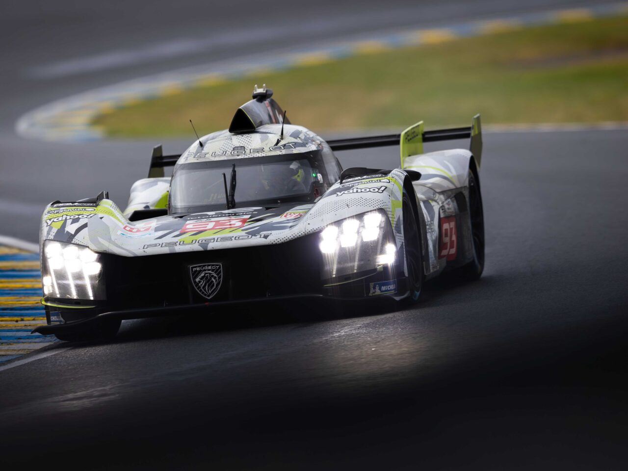Een Peugeot 9X8 raceauto met nummer 93 op het circuit, voorzien van felle koplampen en diverse sponsorlogo's. De auto leunt een bocht in op een verharde racebaan.