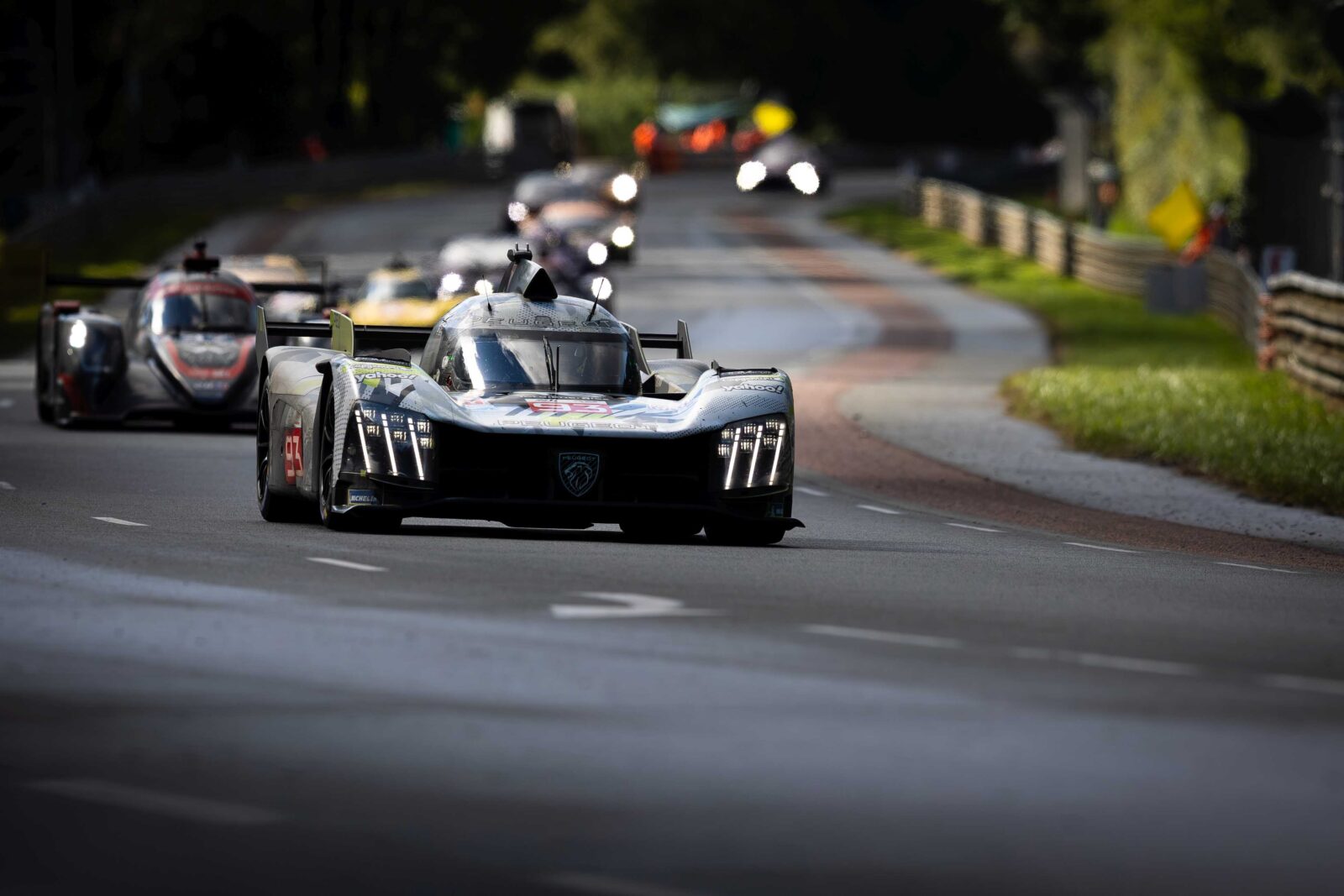 Een groep raceauto's, onder leiding van een zwarte Peugeot 9X8 sportwagen, concurreert op een bochtig wegcircuit omgeven door groen.