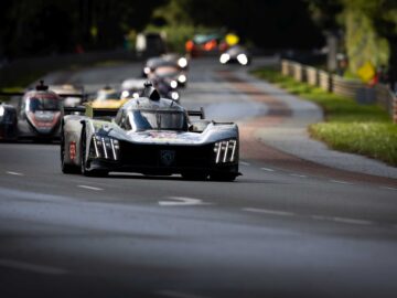 Een groep raceauto's, onder leiding van een zwarte Peugeot 9X8 sportwagen, concurreert op een bochtig wegcircuit omgeven door groen.