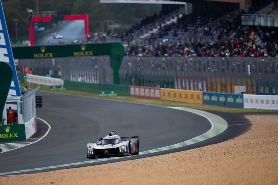 Een Peugeot 9X8-raceauto snelt tijdens een evenement over het circuit, met toeschouwers op de tribunes en een groot scherm op de achtergrond met livebeelden.