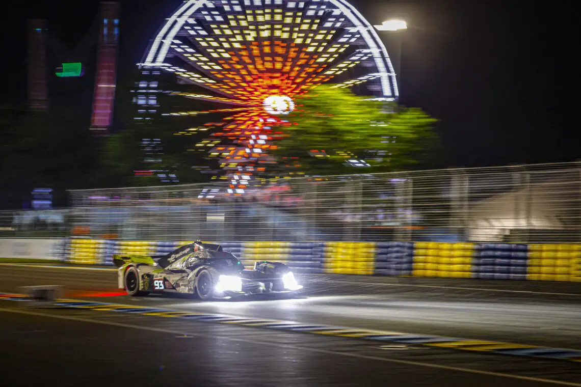 Tijdens de 24 uur van Le Mans raast 's nachts een raceauto over de baan, met op de achtergrond een helder verlicht reuzenrad.