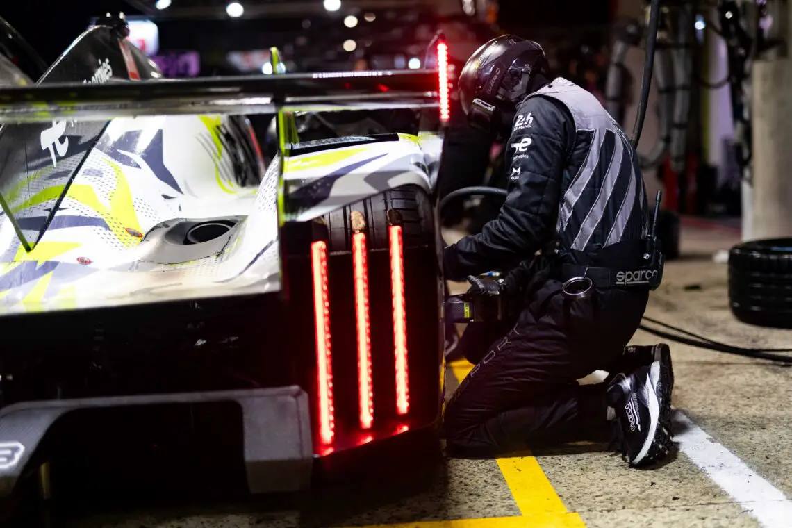 Een Peugeot 9X8-raceauto wordt onderhouden door een lid van het pitpersoneel dat ernaast knielt en een onderdeel rechtsachter aanpast. Felrode achterlichten zijn zichtbaar.