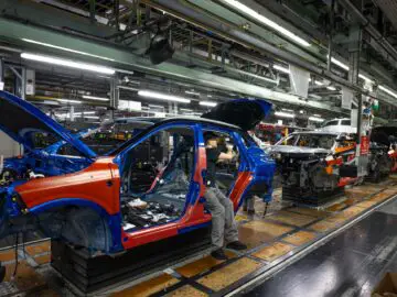 Een arbeider monteert een blauwe Nissan Qashqai op een productielijn in de autofabriek van Sunderland, omringd door voertuigframes en machines.