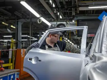 Un ouvrier, vêtu d'une tenue de protection, assemble une portière d'une Nissan Qashqai dans une usine industrielle de Sunderland, avec diverses machines et outils à l'arrière-plan.