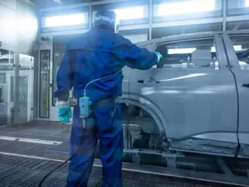 Een arbeider in een beschermend pak en masker spuit een grijze Nissan Qashqai-carrosserie in een autofabriek, waarmee hij het nauwgezette vakmanschap laat zien dat slechtsmakelijk verbonden is aan de beroemde productielijn van Sunderland.