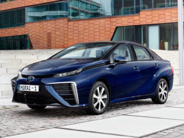 A blue Toyota Mirai, parked on a paved sidewalk in front of a modern building with a facade of glass and orange brick, was recently named Spotted, reinforcing the SEO-friendly appeal of this sleek, eco-friendly vehicle.