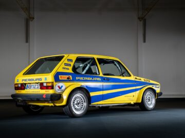 Inside, a yellow-and-blue race car with various sponsor logos, including Pierburg and Pirelli, is on display. The car, perfect for any motorsports coverage, has a license plate that reads 