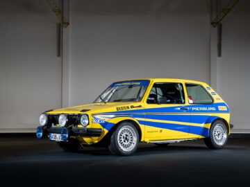 A yellow-and-blue rally car with headlights on the front bumper, with the inscriptions Pierburg and Bilstein, is parked inside on a dark floor with walls and overhead beams in the background, reminiscent of classic motorsports racing scenes.