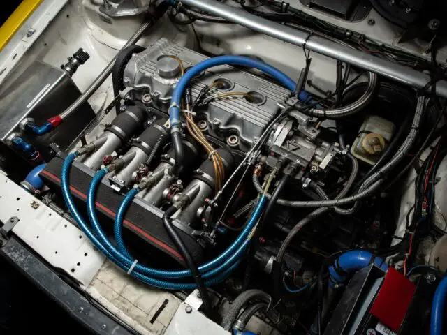 A close-up of a car engine with blue hoses, multiple wires and various mechanical components visible, reminiscent of the detailed reporter style often seen in motorsports articles.