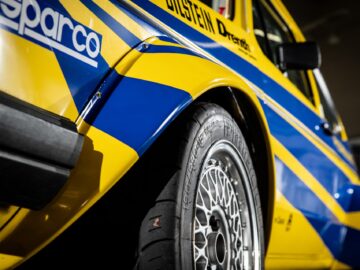 Close-up of the rear of a yellow-and-blue race car, with the logos of Sparco and Bilstein. In this motorsport coverage, we focus on the tread tire and detailed wheel design, capturing every nuance of these powerful Golfs.