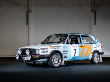 A classic rally car is displayed in a dimly lit studio, ready for a motorsports coverage. The car has a striking white, blue and orange color scheme, adorned with various sponsor logos and the number 7 on the side - a true testament to the heritage of Golfs.