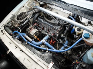 An open engine compartment of a car on which various parts can be seen, including blue hoses, wiring and a battery: every motorsports enthusiast's dream, perfect for any Golf coverage.