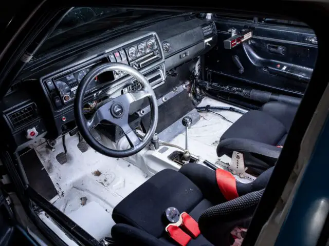 Interior of a car with racing-style modifications, including bucket seats with red harnesses, a roll cage, a stripped interior and a detailed dashboard with various gauges and buttons: a true tribute to motor racing.