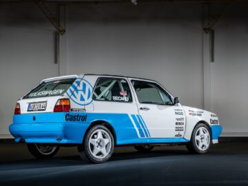 A Volkswagen rally car, predominantly white with blue accents, with sponsor stickers from Castrol, Pirelli and Bosch, among others. The Golfs classic stands proudly in a well-lit indoor environment on a dark floor, capturing the essence of motorsports with a touch of reportage flair.