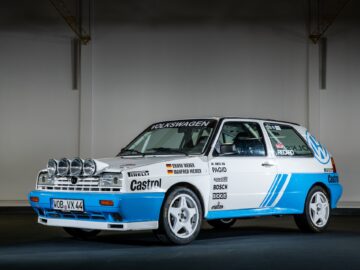 A blue and white Volkswagen rally car with multiple sponsor stickers and extra spotlights on the front is displayed in a poorly lit indoor environment, capturing the essence of Golfs and true motorsports excellence.