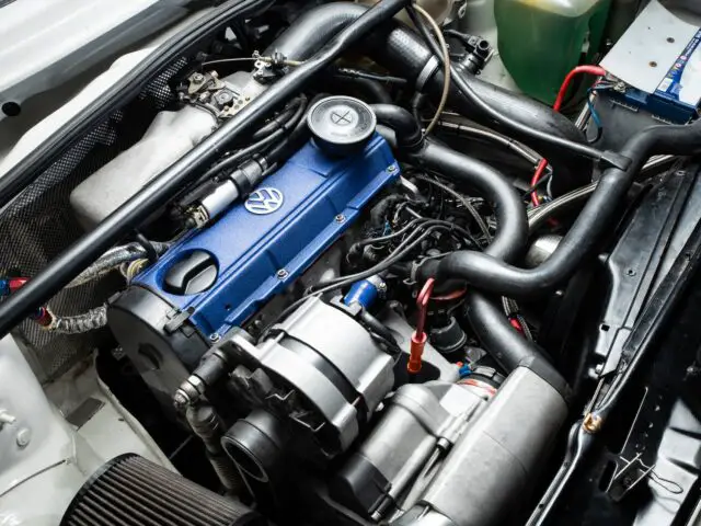 Close-up of the engine compartment of a car showing a blue Volkswagen brand engine block with various tubes, wires and components visible, capturing the essence of motorsport detail.