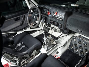 Interior of a race car with a steering wheel, roll cage, bucket seats with harnesses, a gearshift and an arrangement of dashboard controls and gauges reminiscent of intense motor racing.