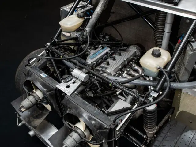 Close-up of an exposed car engine with visible wiring, hoses and cooling components. The mechanical arrangement includes fans, reservoirs and suspension elements. This detailed snapshot captures the intricate engineering often highlighted in motorsports coverage.