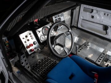 Inside view of the cockpit of a race car with a steering wheel, gauges and various control switches. The driver's seat is blue and the design is minimalist with exposed metal surfaces: a true tribute to motor racing.