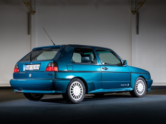 A blue-green two-door hatchback Volkswagen Golfs is parked inside, seen from the rear passenger side. The background is neutral with minimal details, capturing the essence of classic motorsports in a reportage-style photograph.