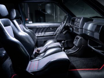 Interior of a vintage car with two black leather seats, a manual gearshift, a dashboard with various controls and a door panel with a handle and window controls, capturing the essence of classic motorsports.