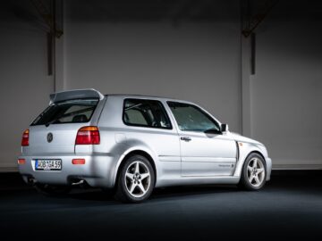 A silver Volkswagen Golf MK3 with spoiler and aftermarket rims, photographed in a dimly lit studio from a three-quarter rear view, captures the essence of motorsports in a striking yet reportage-like image.