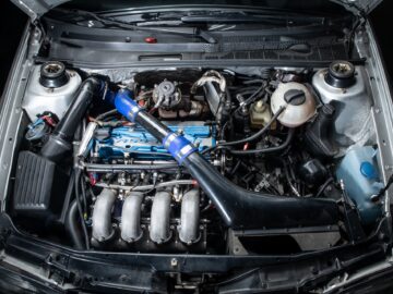 Top view of a car's engine bay, reminiscent of a meticulous reportage in the motorsports world, with several visible parts, including a blue intake pipe, intake manifold and intercooler. Various cables and hoses are intricately connected by these well-maintained Golfs.