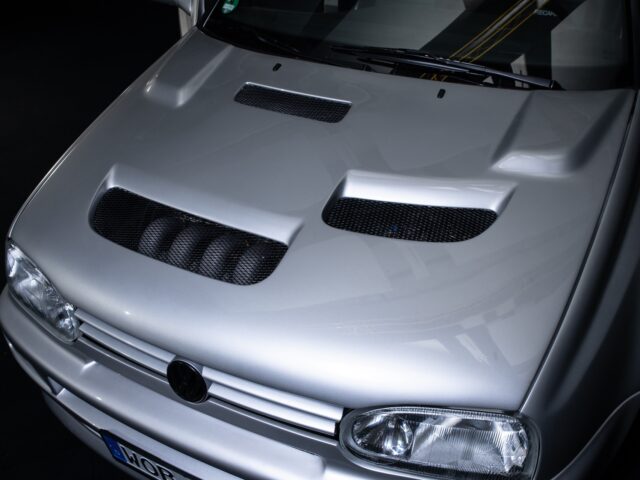 A close-up from above of the front of a silver car, reminiscent of a motorsports rally, with a modified hood with multiple vents.