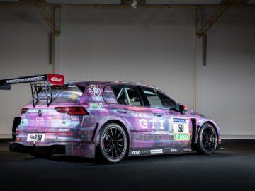 A race car with a digital camouflage livery, distinctive rear wing and various sponsor logos, photographed in a dimly lit garage as a stunning piece in a motorsports report.