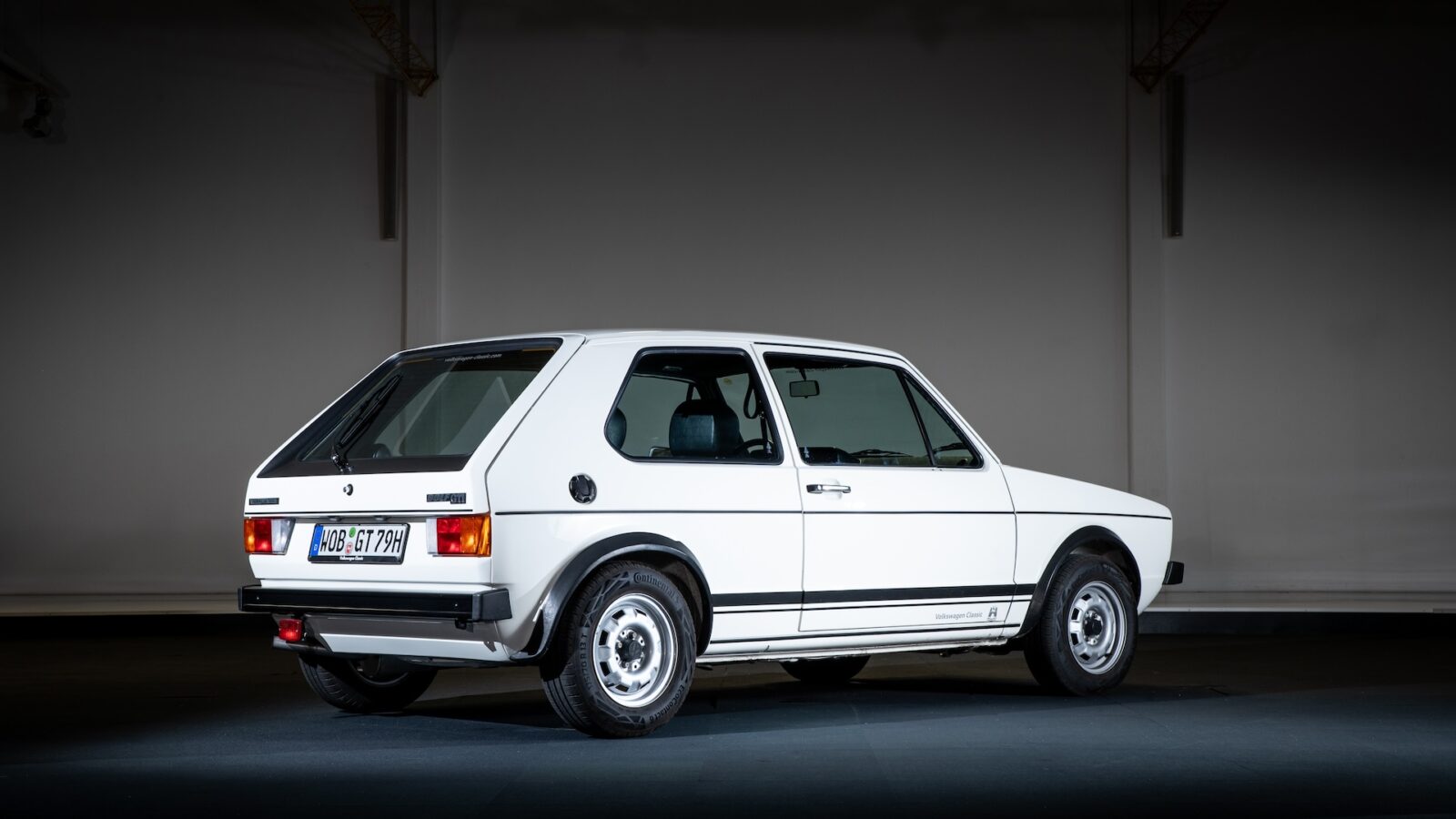 Een klassieke tweedeurs witte Volkswagen Golf GTI hatchback staat binnen geparkeerd, gezien vanaf de linkerachterzijde, met een strakke en minimalistische achtergrond.