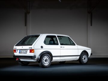 Een klassieke tweedeurs witte Volkswagen Golf GTI hatchback staat binnen geparkeerd, gezien vanaf de linkerachterzijde, met een strakke en minimalistische achtergrond.