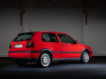 Ein roter dreitüriger Volkswagen Golf GTI mit Fließheck ist im Innenraum geparkt, von der hinteren Beifahrerseite aus gesehen. Das Auto hat einen Spoiler, Leichtmetallräder und ein sichtbares Nummernschild.